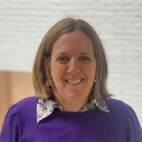Jenny is smiling, wearing a deep purple jumper and earrings that are just visible through her shoulder-length brunette hair.