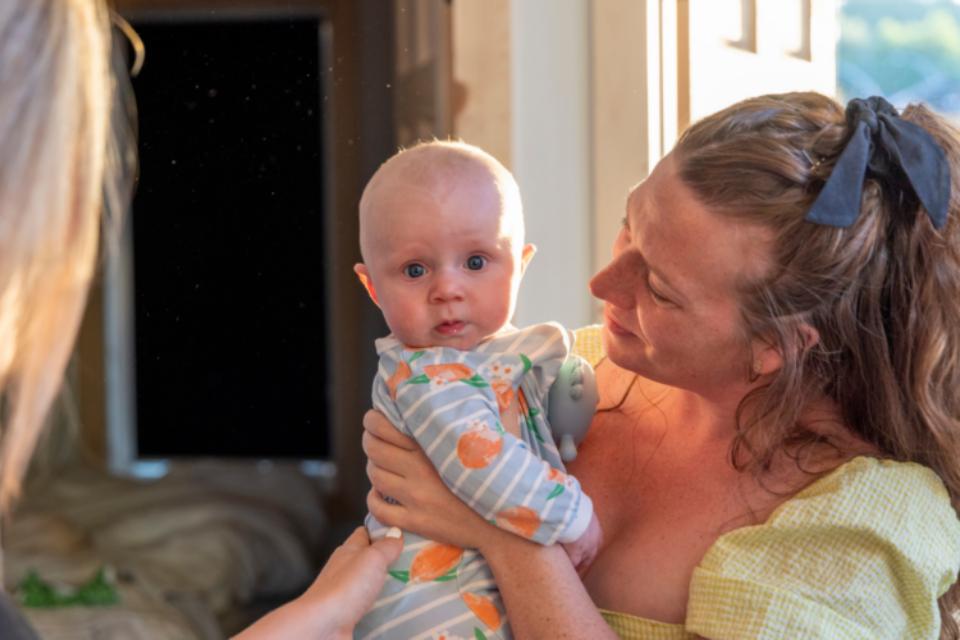 A mum with red, curly hair and fair skin is holding her baby who is looking directly at us. The baby is also fair skinned and has a stripy baby grow on.