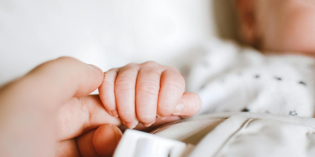 A fair baby's hand wraps around their parent's index finger, they are on white bedding