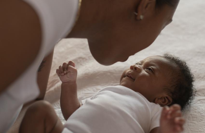 An infant African boy lays on a bed looking up at his mother. The young mother is leaning in close to his face as she looks down at him tenderly. They are both dressed casually.