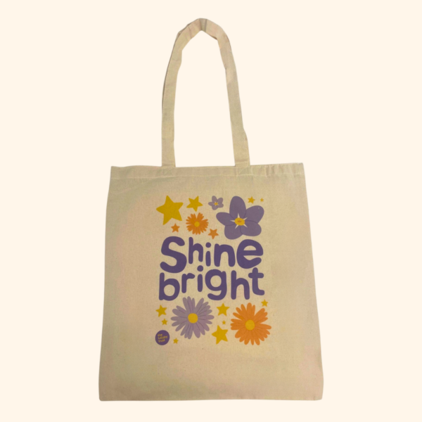 A beige tote bag which says 'Shine bright' in purple bubble letters in the middle. Around the text are purple and orange flowers, yellow stars and a small Lullaby Trust logo in the bottom left