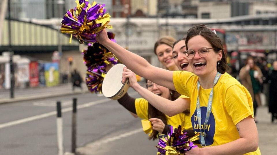 People standing alongside a road cheer runners going past. They are wearing yellow Lullaby Trust t-shirts and waving purple and gold pom poms. The woman at the front is white, with brown hair and glasses.