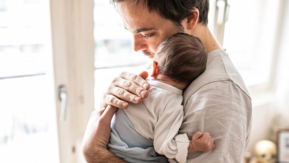 A young baby is resting against the shoulder of their father, who is pressing his head into the baby's and patting their back.