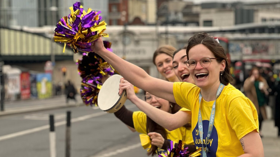 Four people in Lullaby Trust t-shirts cheer along the side of a road. The woman at the front waves a purple and gold pom-pom