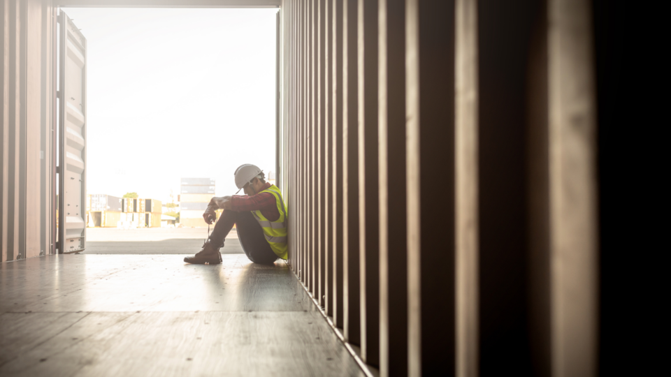 A man is slumped over in a shipping container. He is seemingly at work as wears protective gear.