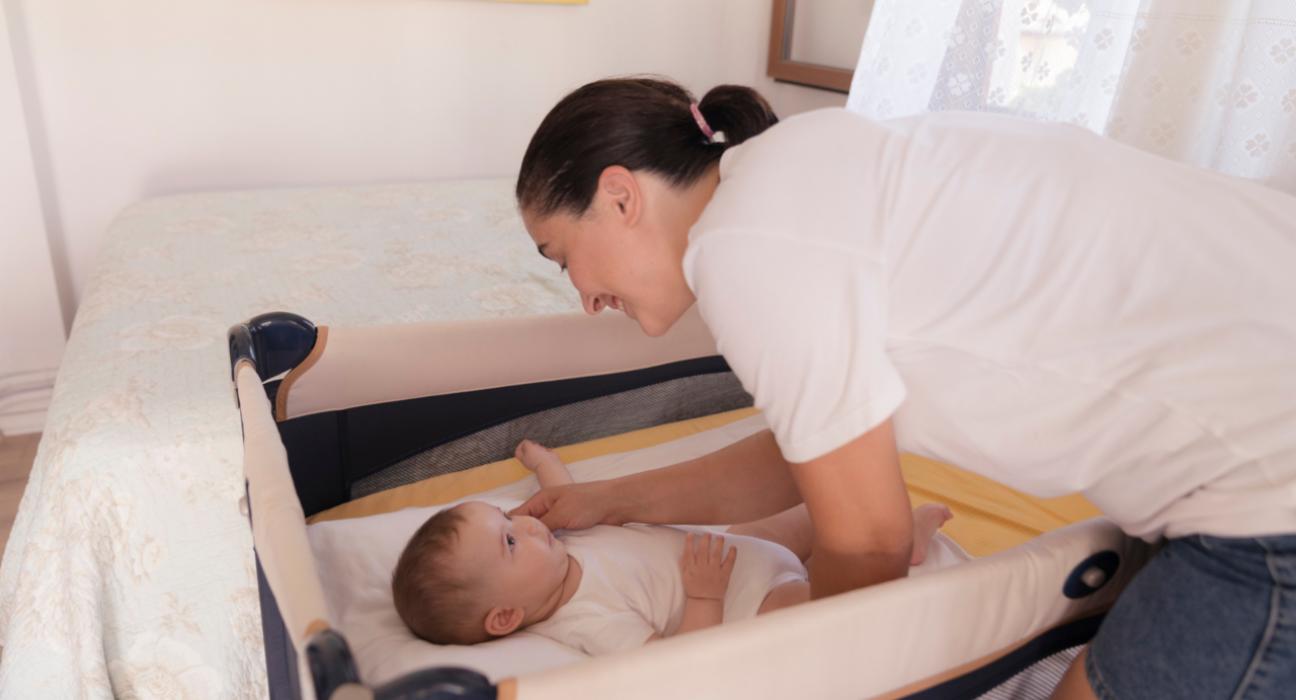 A mum is smiling at her baby who is in a travel cot. She is picking her baby up, and her baby is looking directly at her.