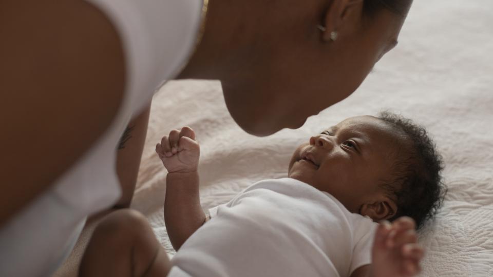 An infant African boy lays on a bed looking up at his mother. The young mother is leaning in close to his face as she looks down at him tenderly. They are both dressed casually.