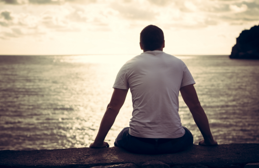 Man is sitting looking out at the sea. He has a muscular silhouette, shaved head and is slumped slightly.