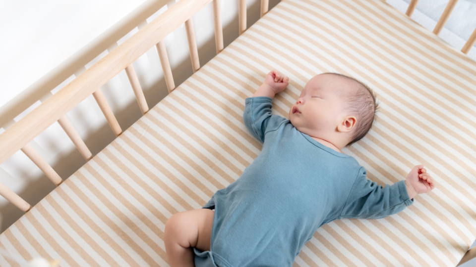 A baby in a blue baby grow is sleeping on their back in a wooden cot with a firm mattress.