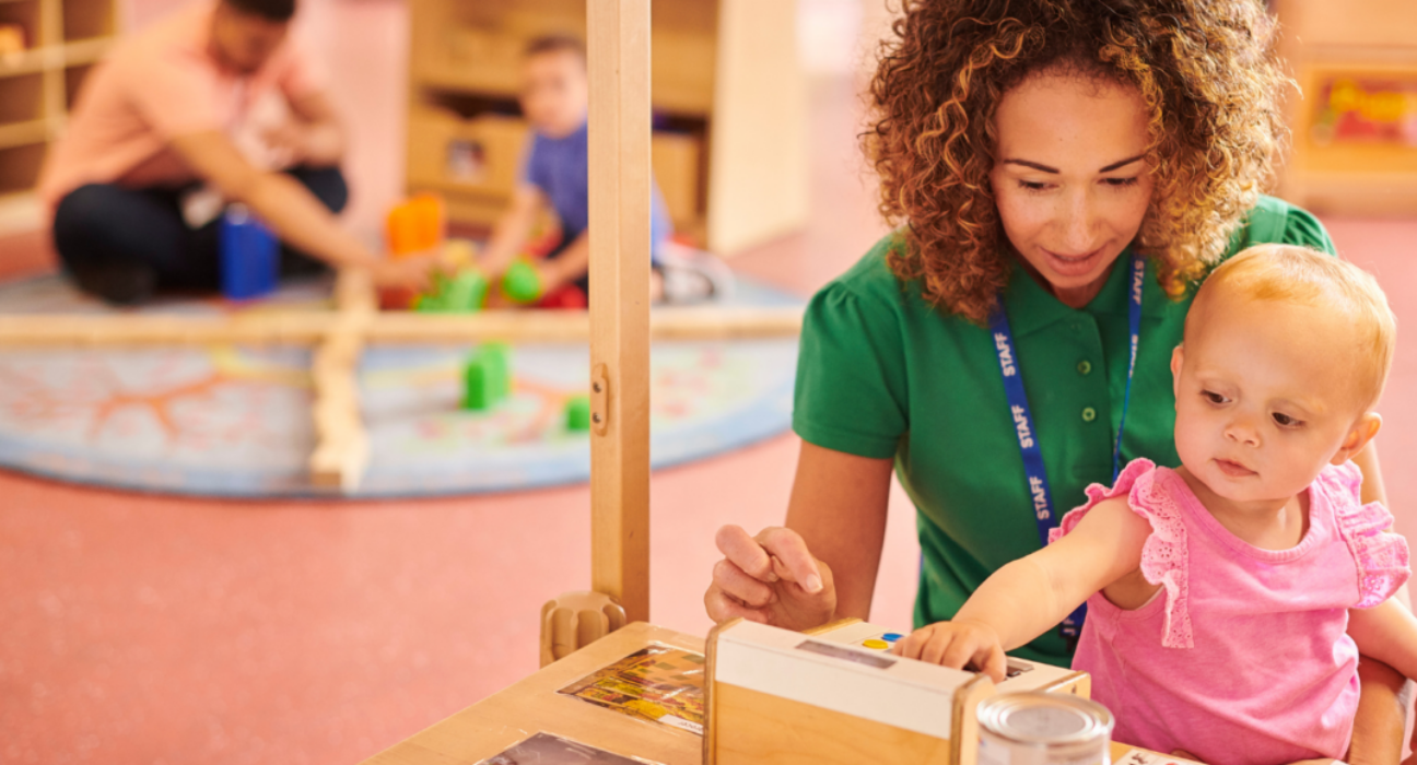 Early years professional sitting on the floor in a nursery with a little girl on her lap, playing with toys.