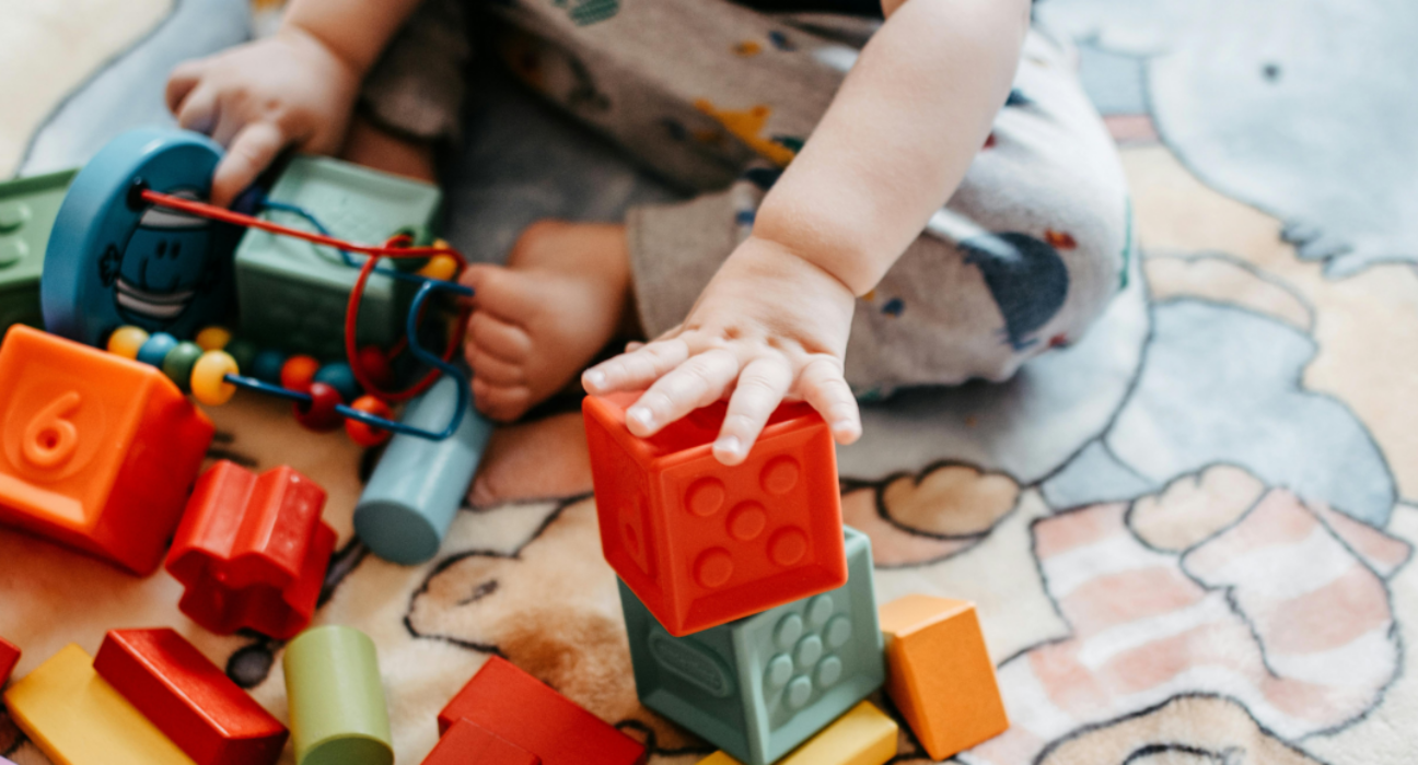 A close up of a baby's hand playing with some blocks. The baby is sitting, playing with plastic and wooden colourful blocks on a mat.