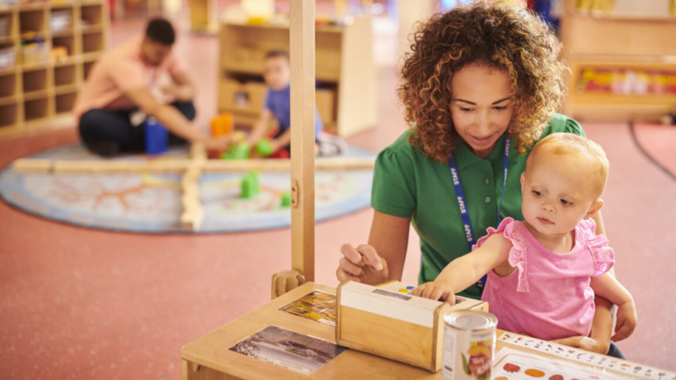 Early years professional sitting on the floor in a nursery with a little girl on her lap, playing with toys.