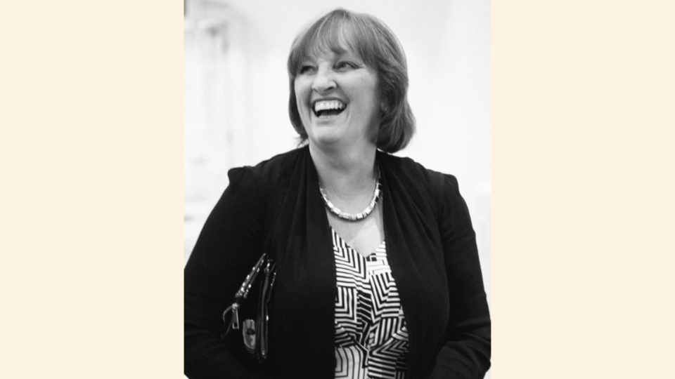 A black and white photo of Bridget. She is dressed formally with a dress, blazer and pearl necklace. Her head is turned slightly to the side and she is mid laugh with a bright smile.