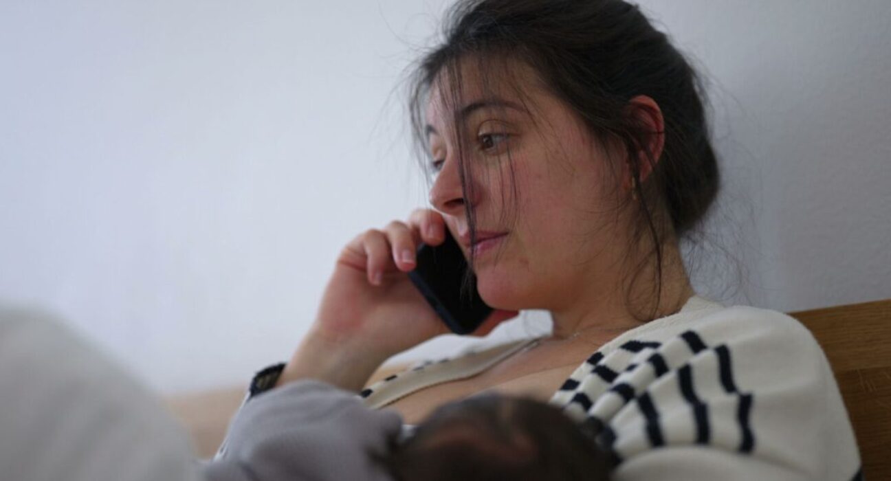 Mother multitasking while her baby sleeps peacefully in her arms, speaking on the phone with a thoughtful expression. the quiet balance between caring for a sleeping newborn and handling daily responsibilities