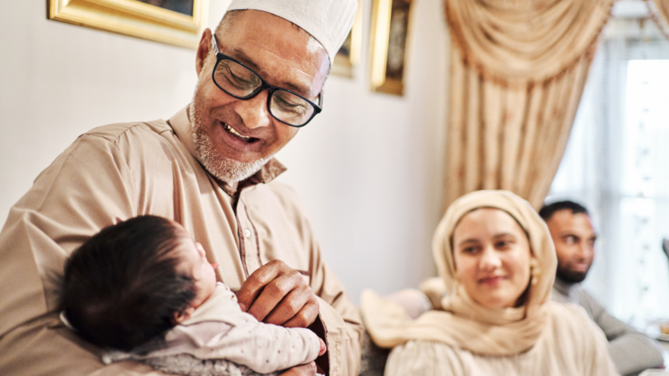 A grandfather is smiling, wears glasses and holding his grandchild, touching his chest with one hand. A woman is blurred out in the distance, wearing a hijab and is grinning, watching on.
