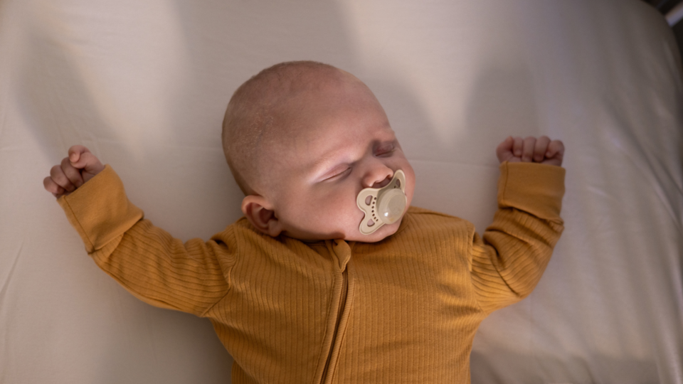 A baby is sleeping with their arms either side of their head. They're wearing yellow, have light skin and a dummy in their mouth.