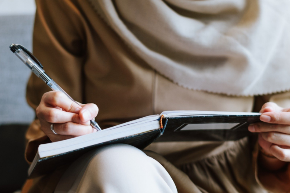 Woman wearing a hijab, close up of her hand writing in a notebook using a pen.