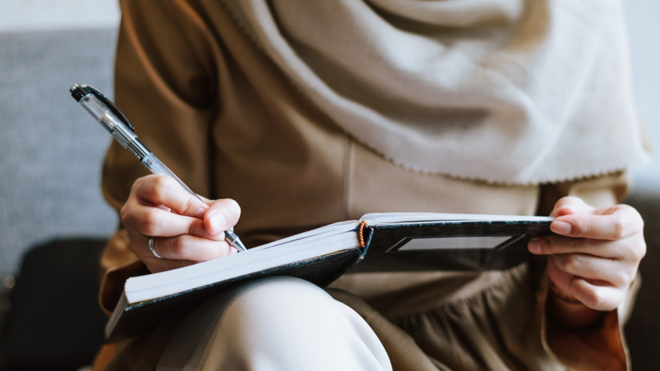 Woman wearing a hijab, close up of her hand writing in a notebook using a pen.