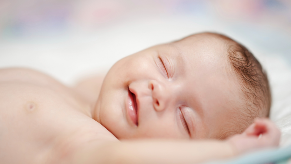 A baby is sleeping and smiling in their sleep. Their arms are either side of their head.