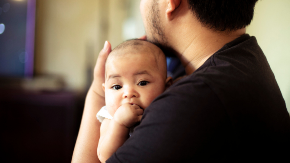 A dad is holding a baby close to his chest. The baby is chewing their hands.