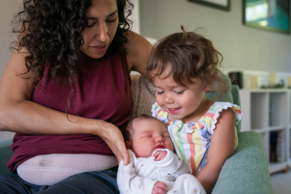 A woman sits on the sofa at home and helps her cute toddler daughter hold her baby brother for the first time. The big sister appears excited and proud.