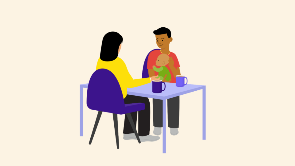 A man holding a baby, sitting at a table opposite another person. There are mugs on the table.