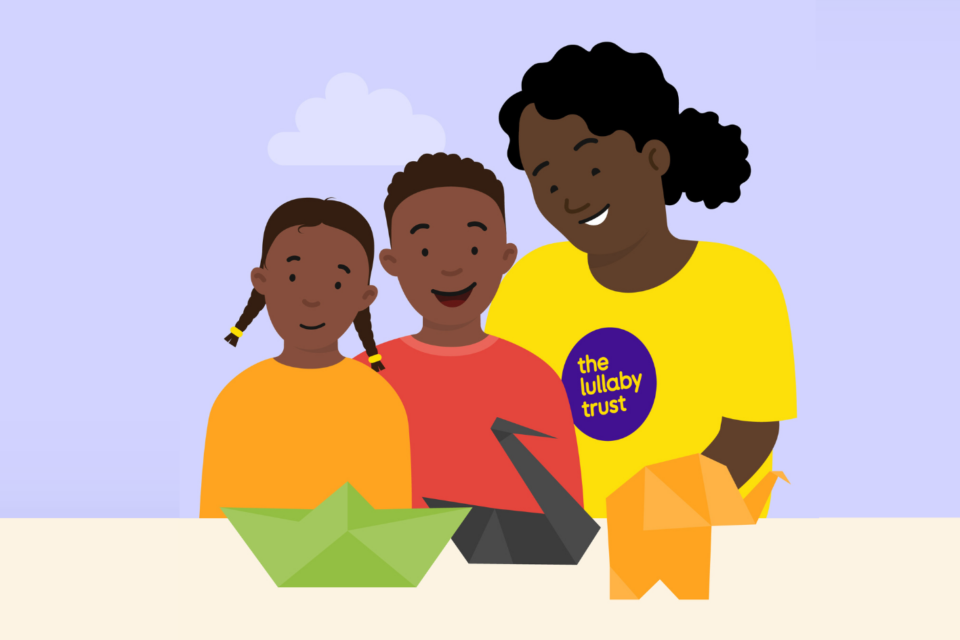 Illustration of a lady wearing a Lullaby Trust t-shirt standing beside two children who are smiling. On the table in front of them are some origami animals.