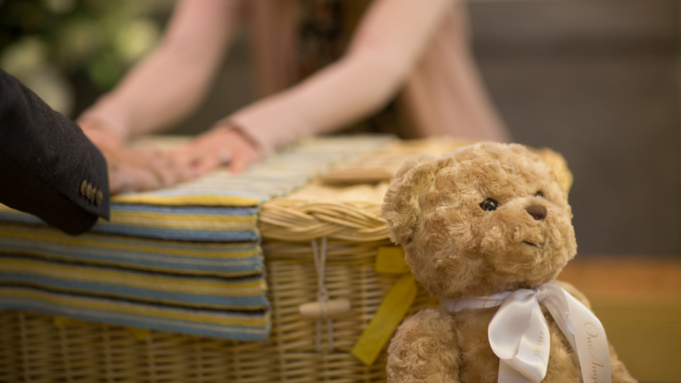 Holding hands over a wicker basket at a funeral, with a teddy bear leaning on it.