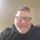 Dan, Trustee, smiling at the camera with a black jumper on and glasses.