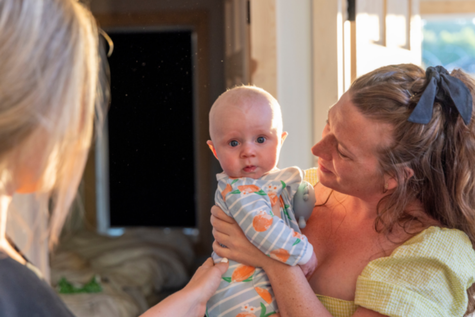 Lady holding a baby, and another lady standing in front of her with her hand on the baby's back.