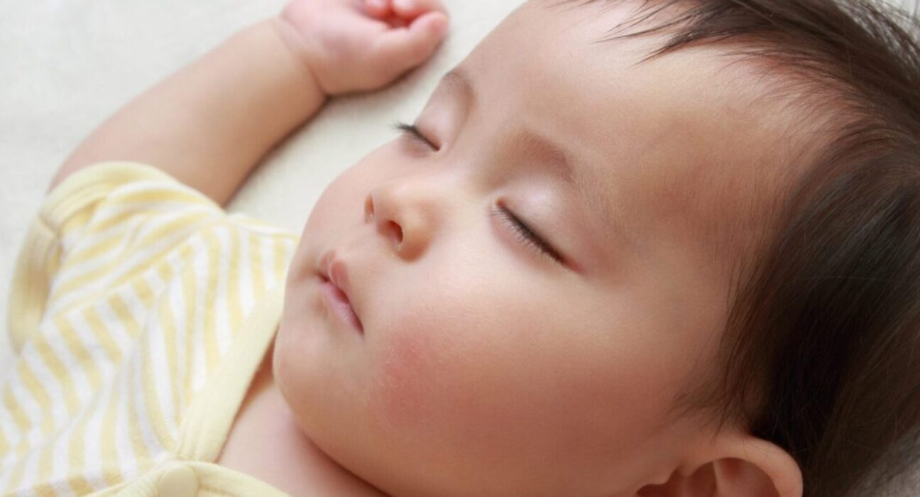 Close up of a baby's face, sleeping on their back and wearing a yellow stripy baby grow.