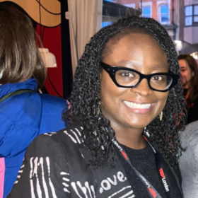 Seyi wearing a black and white top and black glasses, smiling.