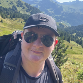 Matt wearing a cap and sunglasses, with a backpack on his back. He is taking a selfie in front of a vast natural landscape.