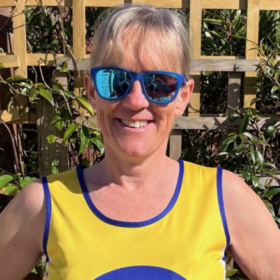 Clare wearing sunglasses and her Lullaby Trust vest, standing outdoors smiling at the camera.