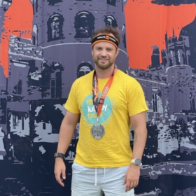 Stefan wearing a Lullaby Trust t-shirt and a medal, standing in front of a big tough mudder backdrop.