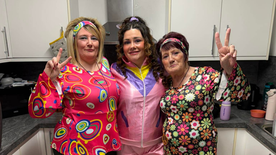 Barbara and her daughters all wearing 70s themed clothes, holding a peace sign up at the camera.