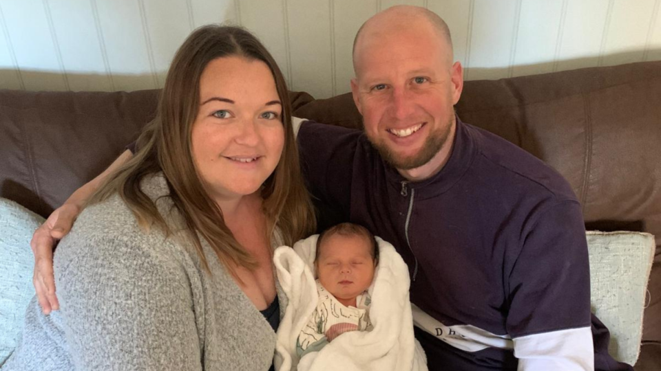 Sarah and Craig with baby Ezrah, sitting on a sofa and smiling at the camera.