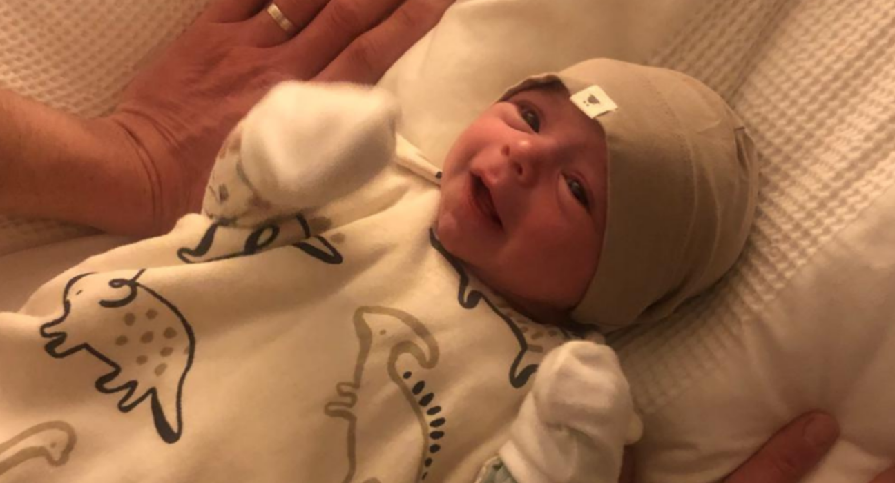 Ezrah lying on a bed with a hat on, smiling. Two adults hands are resting either side of him.