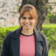 Lalitha wearing a pink top and a jacket standing outside, smiling at the camera.