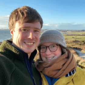 Matt and Laura are taking a selfie in front of green grassy moors. They are dressed warmly and both smiling.