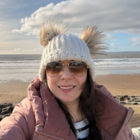 Becky taking a selfie by the sea, wearing a wooly hat and smiling at the camera.