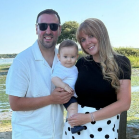 Leah and George standing outdoors, holding baby George and smiling at the camera