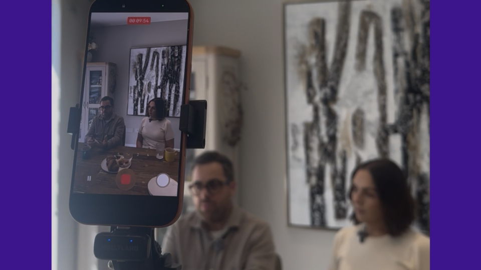 Katie and John Meehan being interviewed behind a mobile phone, with a video recording live. They are sat down at a table.