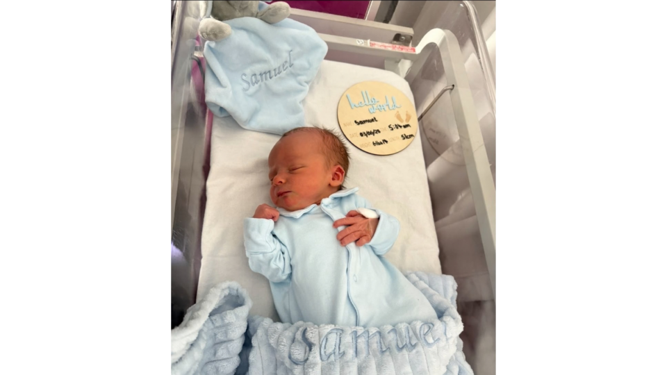 Samuel when he was born. He has a blue blanket with his name on and a blue babygrow. There is a wooden circle saying 'hello world' with his name, weight and length.
