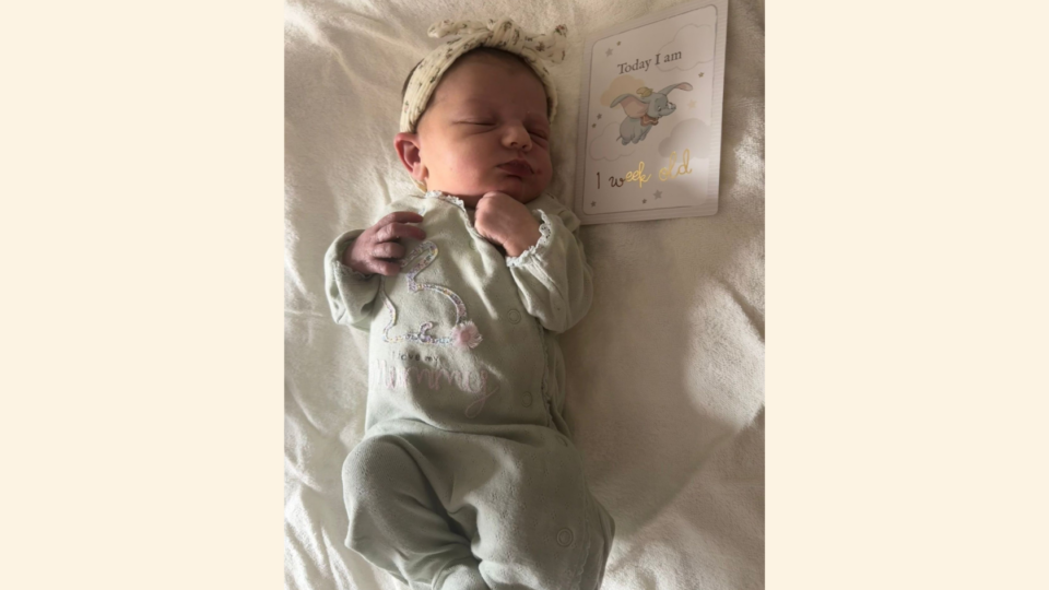 Magnolia lying on bed with a headband on, and a card next to her that says 'today I am 1 week old'