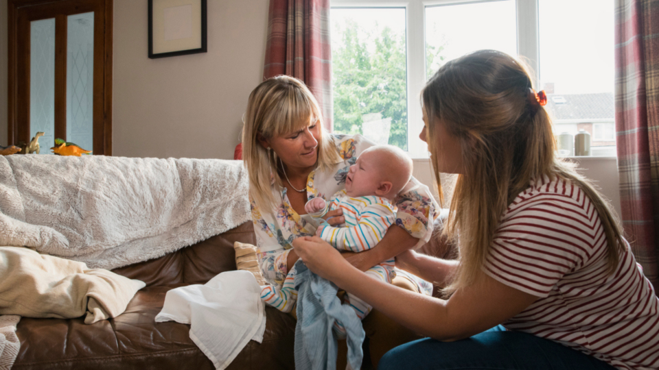 The mother of a baby has just handed the baby over to the grandmother. The baby is crying, both women are looking directly at the baby adoringly.