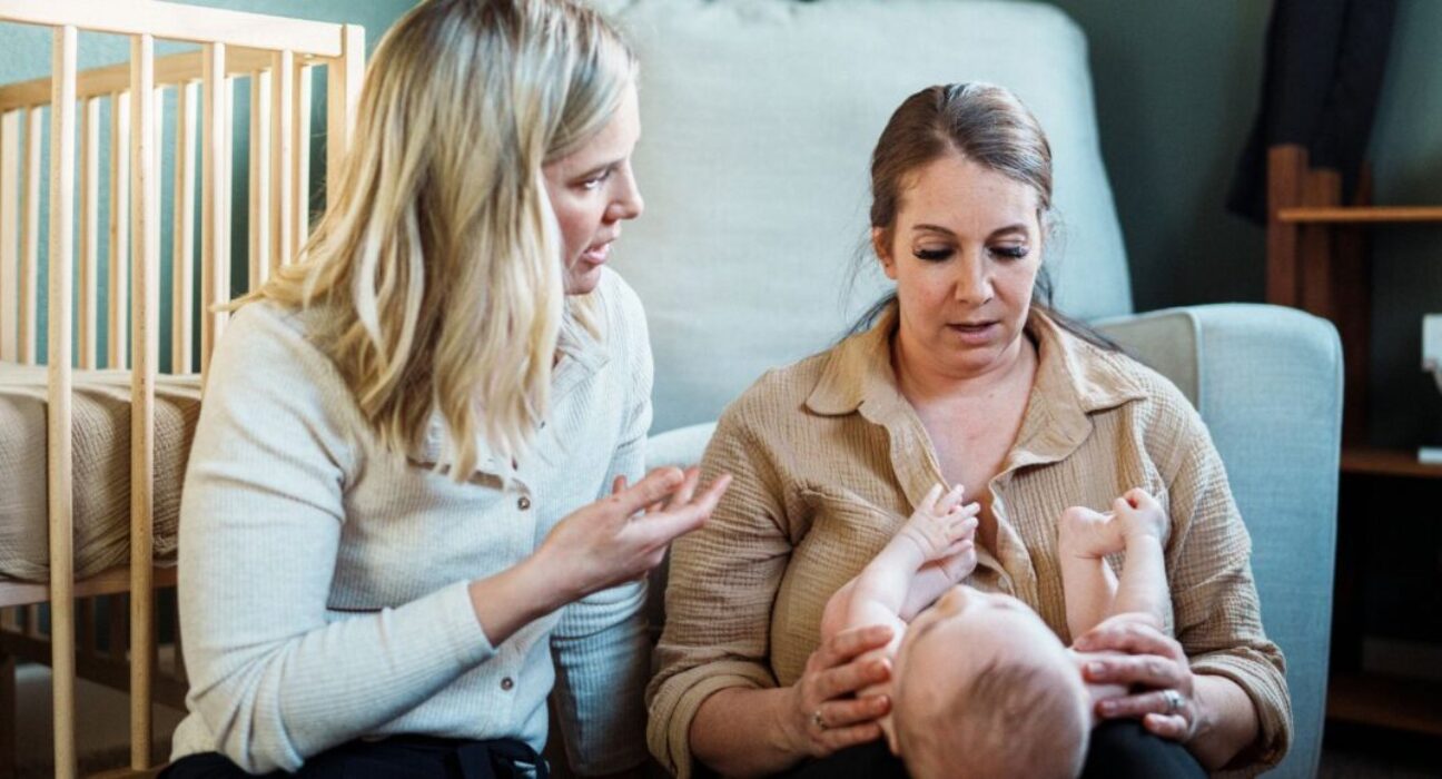 A mum is looking down at her baby in her knee. A health visitor is next to them, talking to the mother. A cot is in the background.