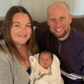 Sarah and Craig with baby Ezrah, sitting on a sofa and smiling at the camera.