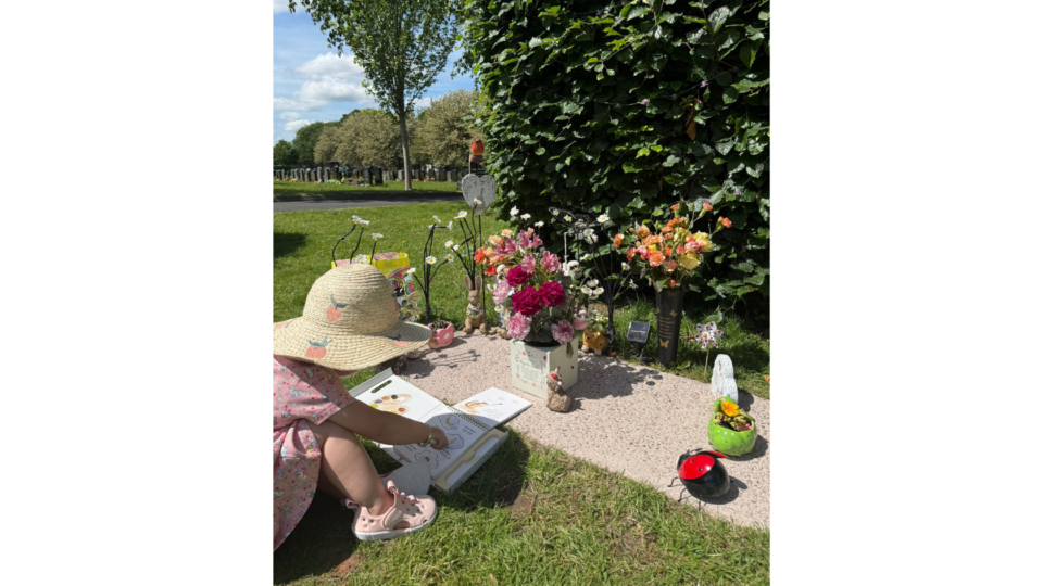 Cordelia at Magnolia's grave, reading a book. There are lots of flowers on the grave.
