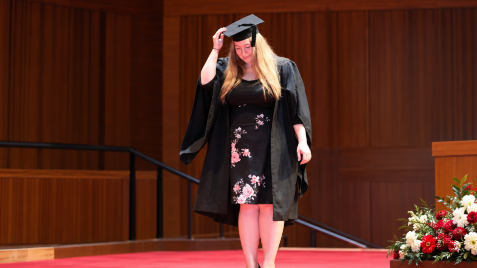 Michaela on stage at graduation, wearing graduation gown and hat and a dress.